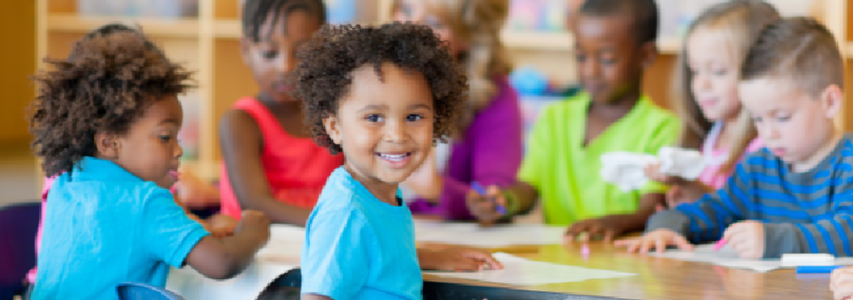 A smiling child sitting at a classroom table with other children and an adult working on papers behind him.
