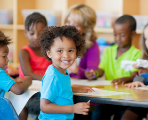 A smiling child sitting at a classroom table with other children and an adult working on papers behind him.
