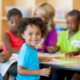 A smiling child sitting at a classroom table with other children and an adult working on papers behind him.