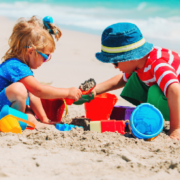 Two children playing with colorful buckets, shovels, and sand toys on a sandy beach near blue ocean waves.