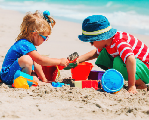 Two children playing with colorful buckets, shovels, and sand toys on a sandy beach near blue ocean waves.