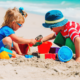 Two children playing with colorful buckets, shovels, and sand toys on a sandy beach near blue ocean waves.