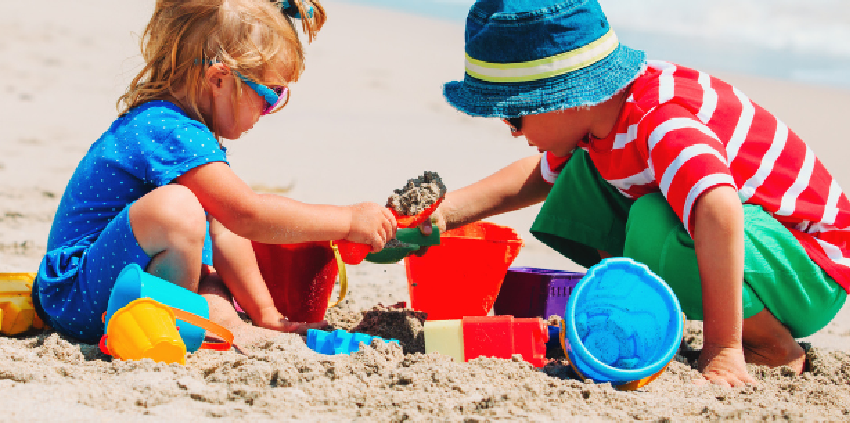 Two children playing with colorful buckets, shovels, and sand toys on a sandy beach near blue ocean waves.