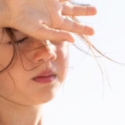 A child shields her eyes from bright summer sunlight while standing outdoors with loose hair blowing across her face.
