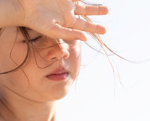 A child shields her eyes from bright summer sunlight while standing outdoors with loose hair blowing across her face.
