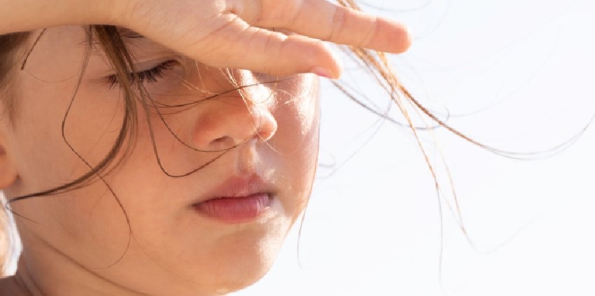 A child shields her eyes from bright summer sunlight while standing outdoors with loose hair blowing across her face.