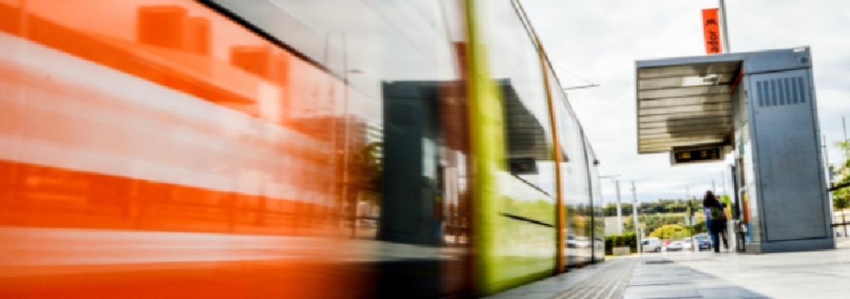 An orange train moving beside a station platform with tactile paving, overhead sign, cloudy sky, and distant people.