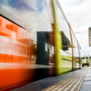 An orange train moving beside a station platform with tactile paving, overhead sign, cloudy sky, and distant people.
