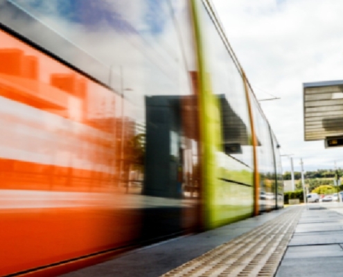 An orange train moving beside a station platform with tactile paving, overhead sign, cloudy sky, and distant people.
