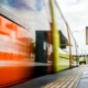 An orange train moving beside a station platform with tactile paving, overhead sign, cloudy sky, and distant people.