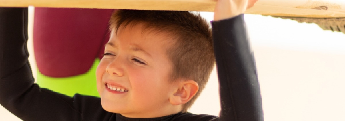 A smiling young boy in a black wetsuit carries a surfboard over his head while spending a sunny day at the beach.