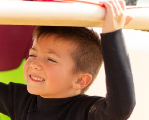 A smiling young boy in a black wetsuit carries a surfboard over his head while spending a sunny day at the beach.