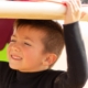 A smiling young boy in a black wetsuit carries a surfboard over his head while spending a sunny day at the beach.