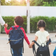 Parent walks three young children carrying backpacks toward school together on a sunny school morning.