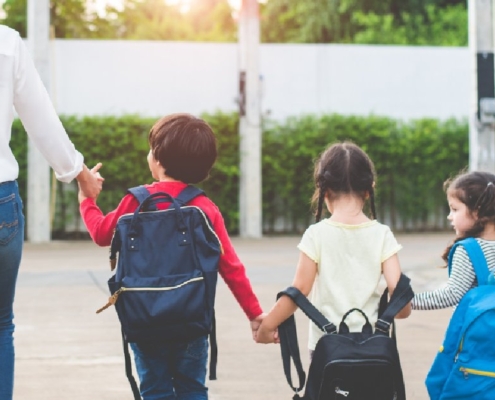 Parent walks three young children carrying backpacks toward school together on a sunny school morning.