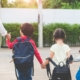 Parent walks three young children carrying backpacks toward school together on a sunny school morning.