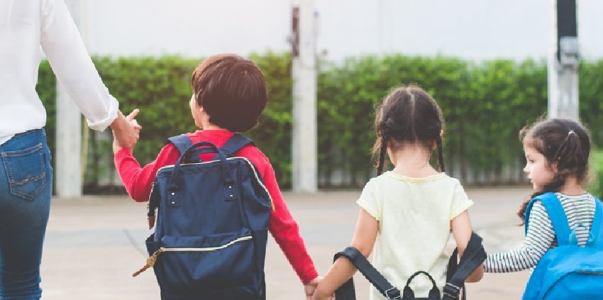 Parent walks three young children carrying backpacks toward school together on a sunny school morning.