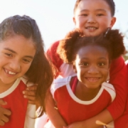 Young athletes in red and white sports jerseys huddle closely together and smile outdoors before a team game.